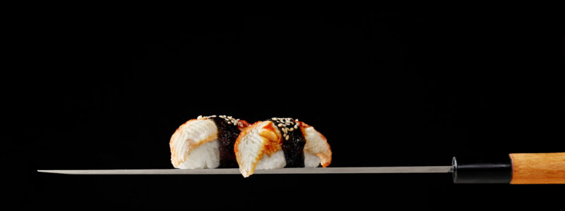 Nigiri sushi topped with grilled eel slices wrapped with nori sprinkled with sesame seeds served on blade of traditional Japanese knife yanagiba isolated on black background. Authentic cuisine