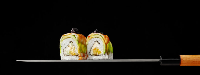 Two uramaki rolls with cream cheese, mango, avocado and shrimps topped with tobiko served on thin stainless steel blade of traditional Japanese kitchen knife on black background